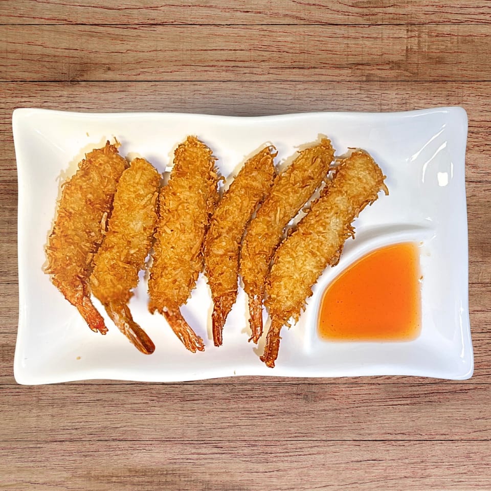 Seven golden-brown coconut shrimp with tails on, served with orange dipping sauce on a white plate.