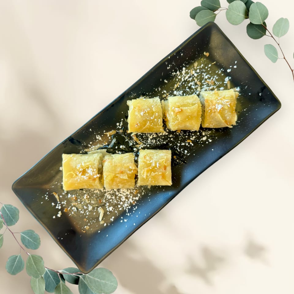 Six pieces of golden-brown fried banana spring rolls, dusted with powdered sugar, on a black rectangular plate.