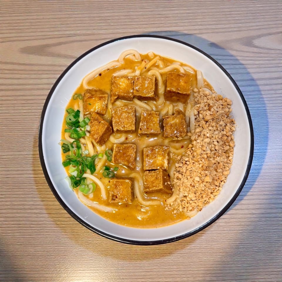 A bowl of udon noodles with tofu, green onions, and crushed peanuts in a rich sauce.