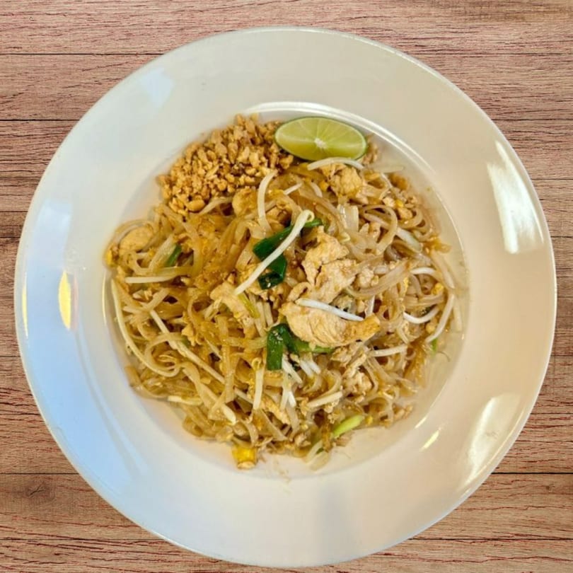A white plate of Pad Thai noodles with chicken, bean sprouts, green onions, crushed peanuts, and a lime wedge.
