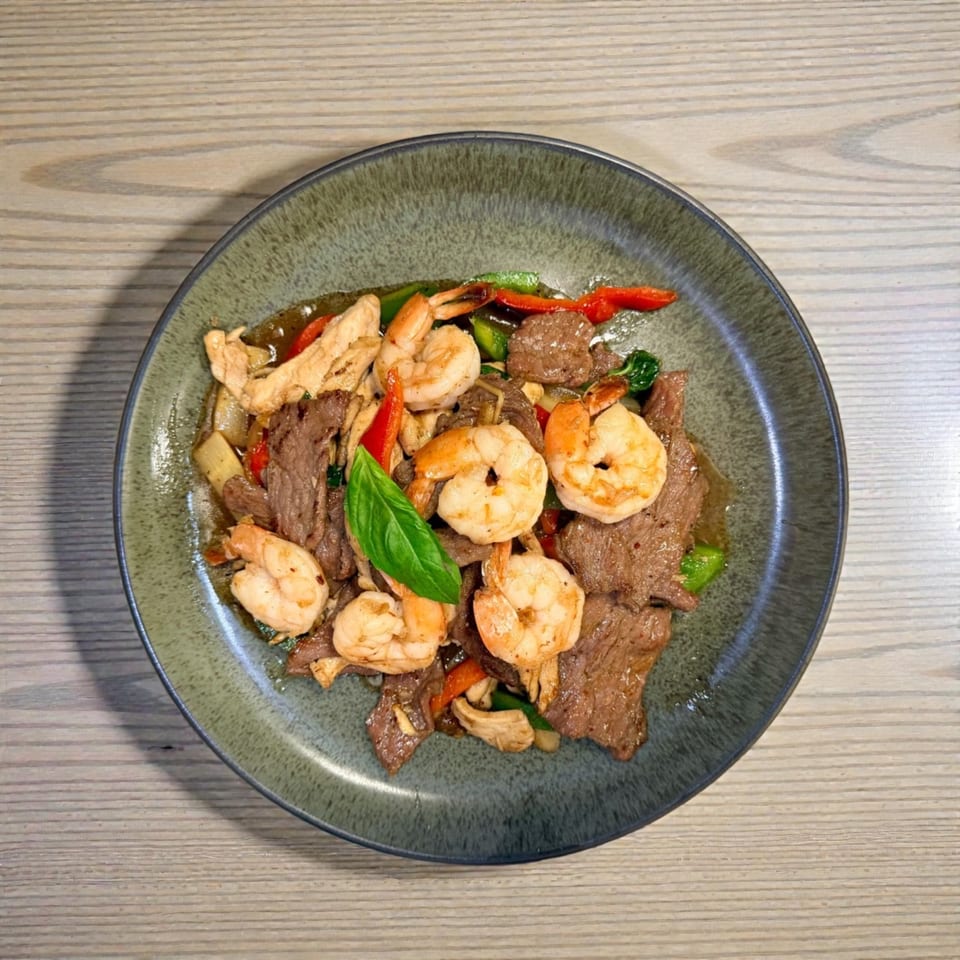 A green plate holds a stir-fry with beef, shrimp, chicken, red peppers, and a basil leaf.