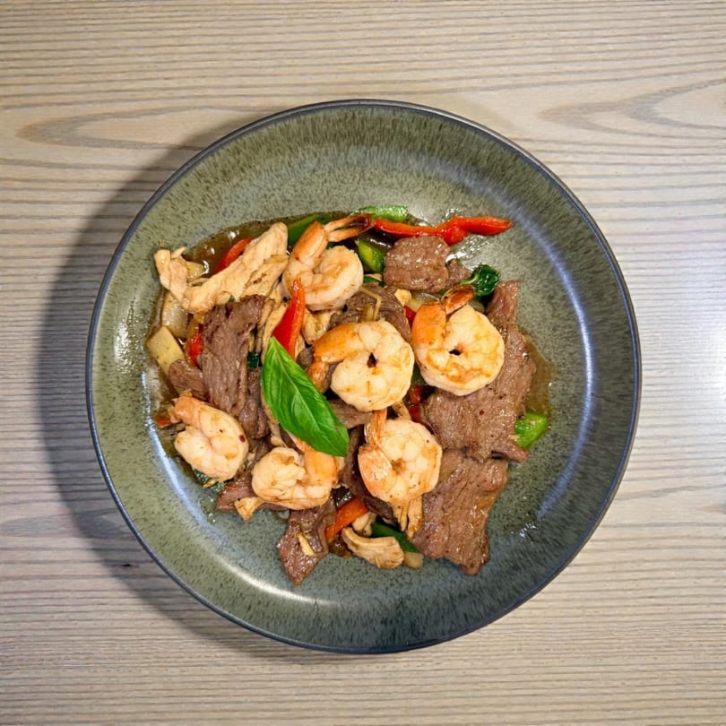 A green plate holds a stir-fry with beef, shrimp, chicken, red peppers, and a basil leaf.