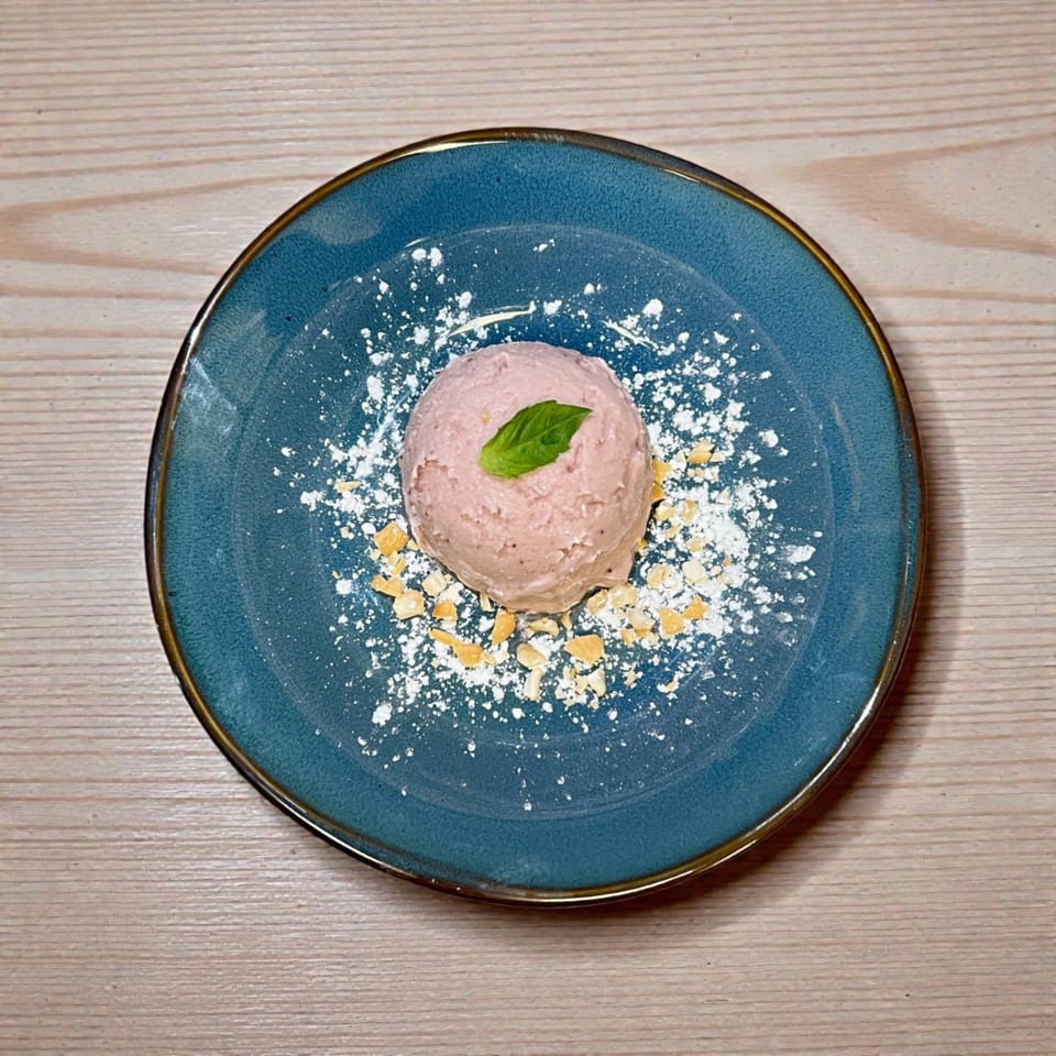 A scoop of pink ice cream with a mint leaf, powdered sugar, and nuts on a blue plate.